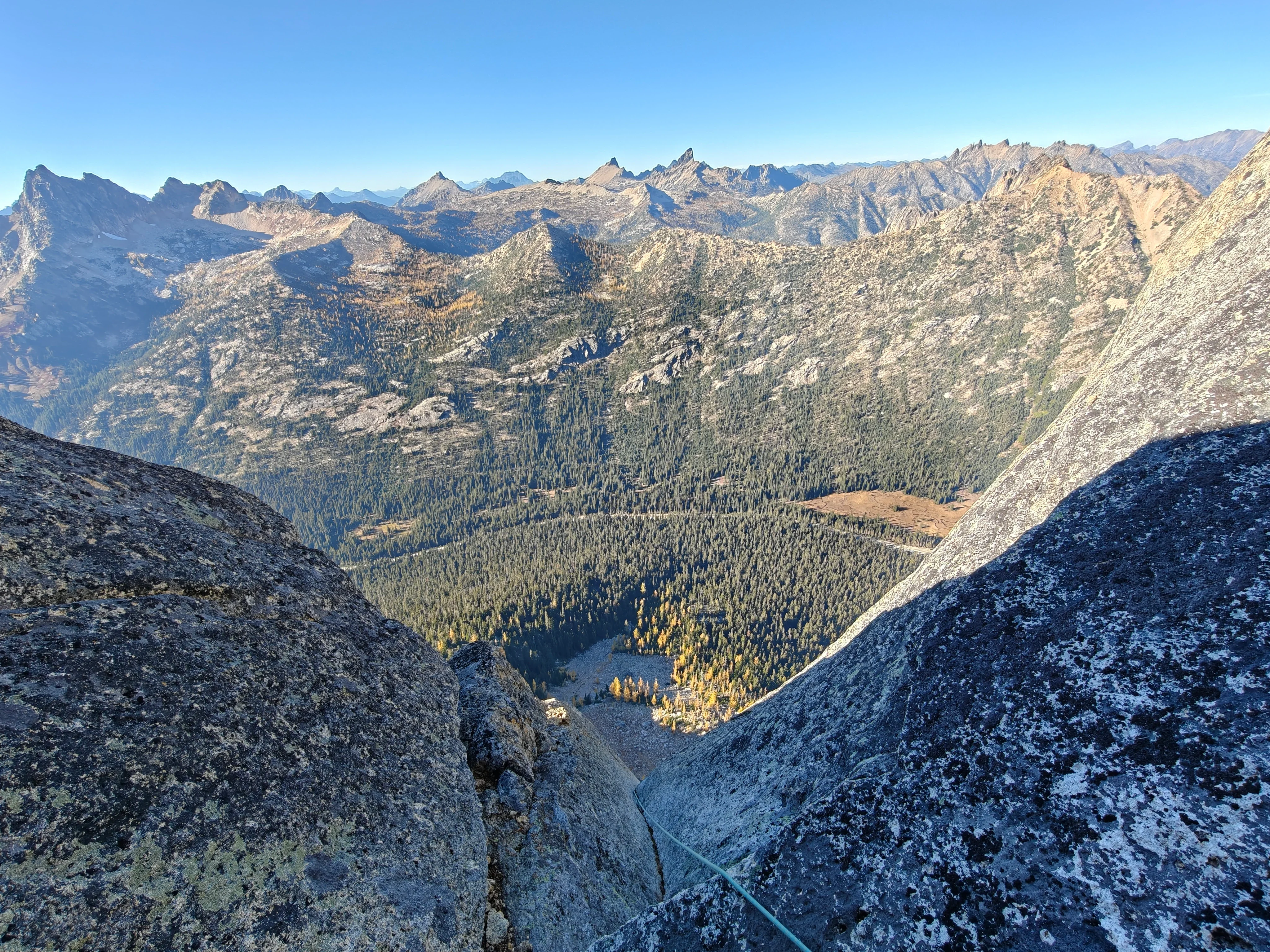 Teal rope disappearing down a granite chimney, with forested valleys and jagged peaks beyond