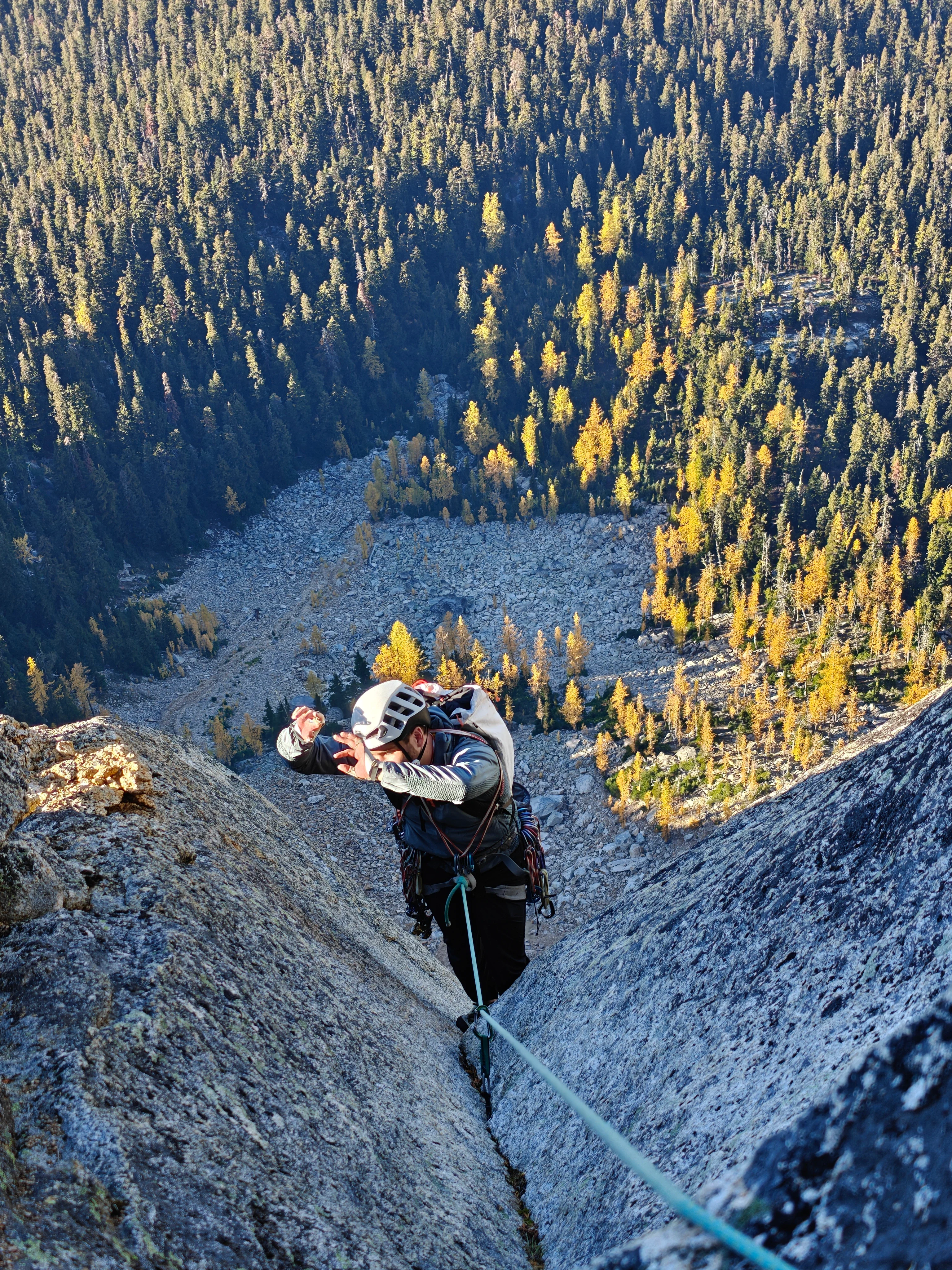 Climber following a crack, shielding their eyes from the sun, with talus and golden larches far below
