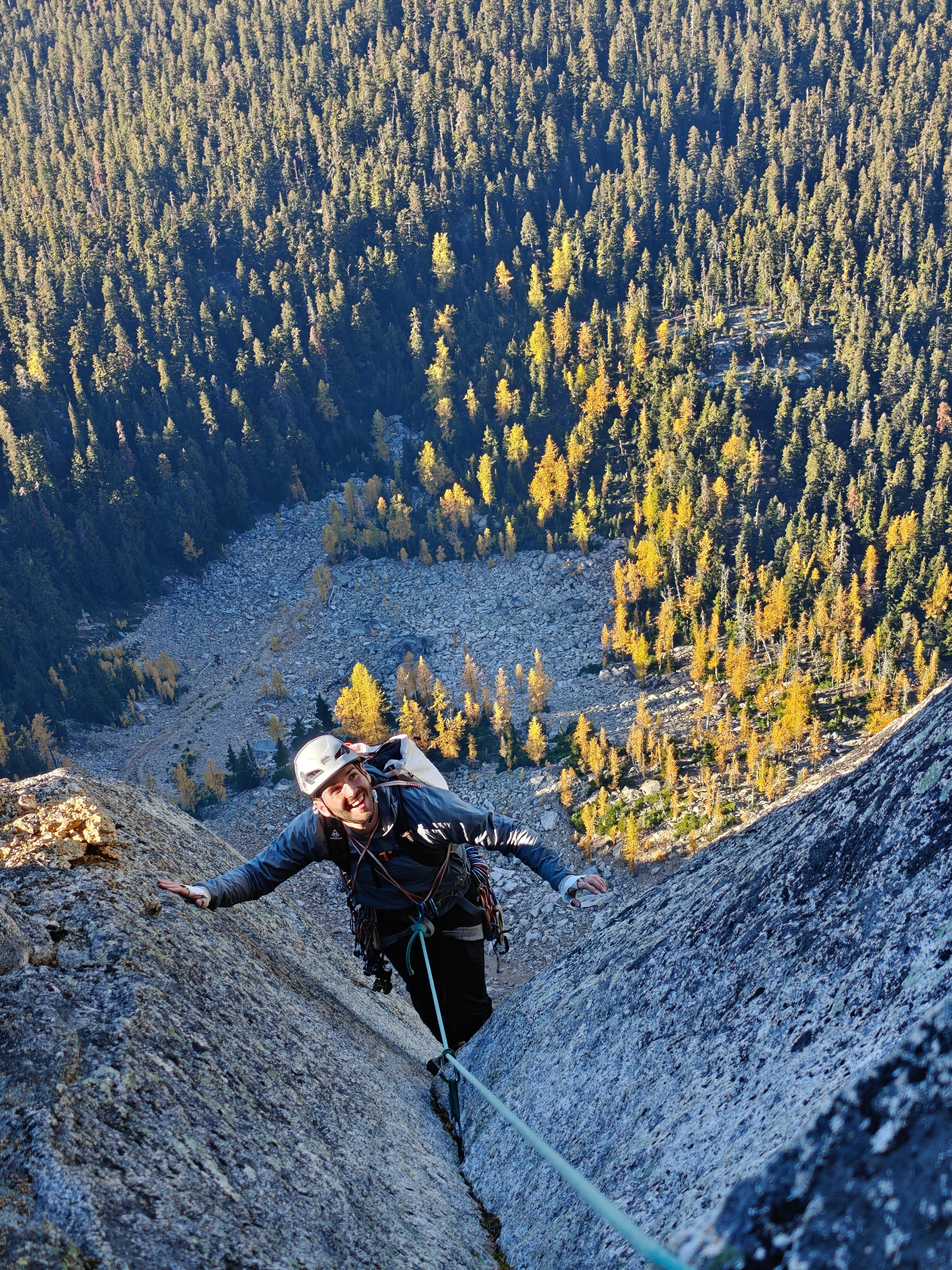 Climber in a white helmet smiling up from a granite crack, golden larches and talus far below
