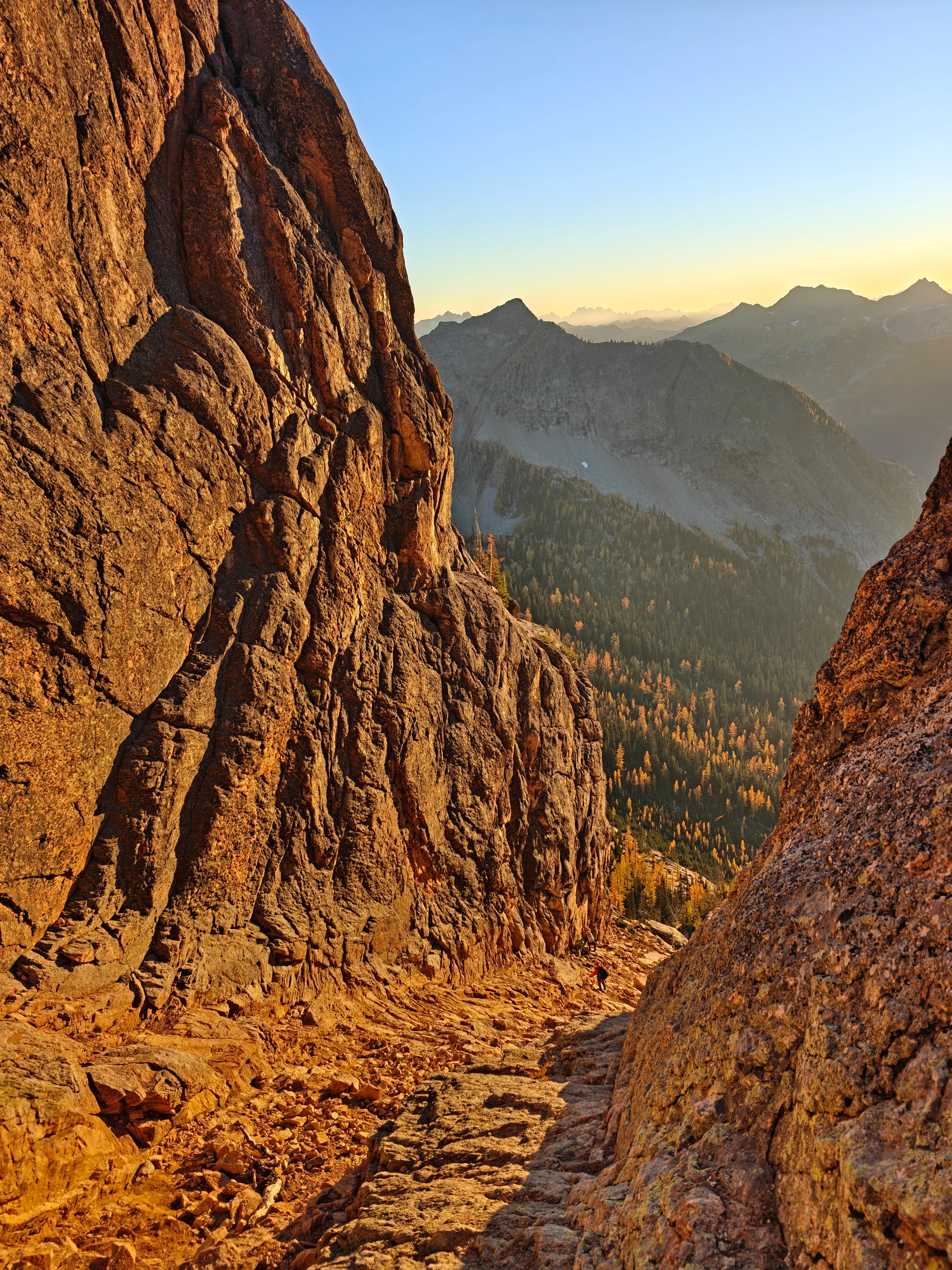 Descent gully lit orange at sunset between towering rock walls, a small climber partway down and larch-covered valley beyond