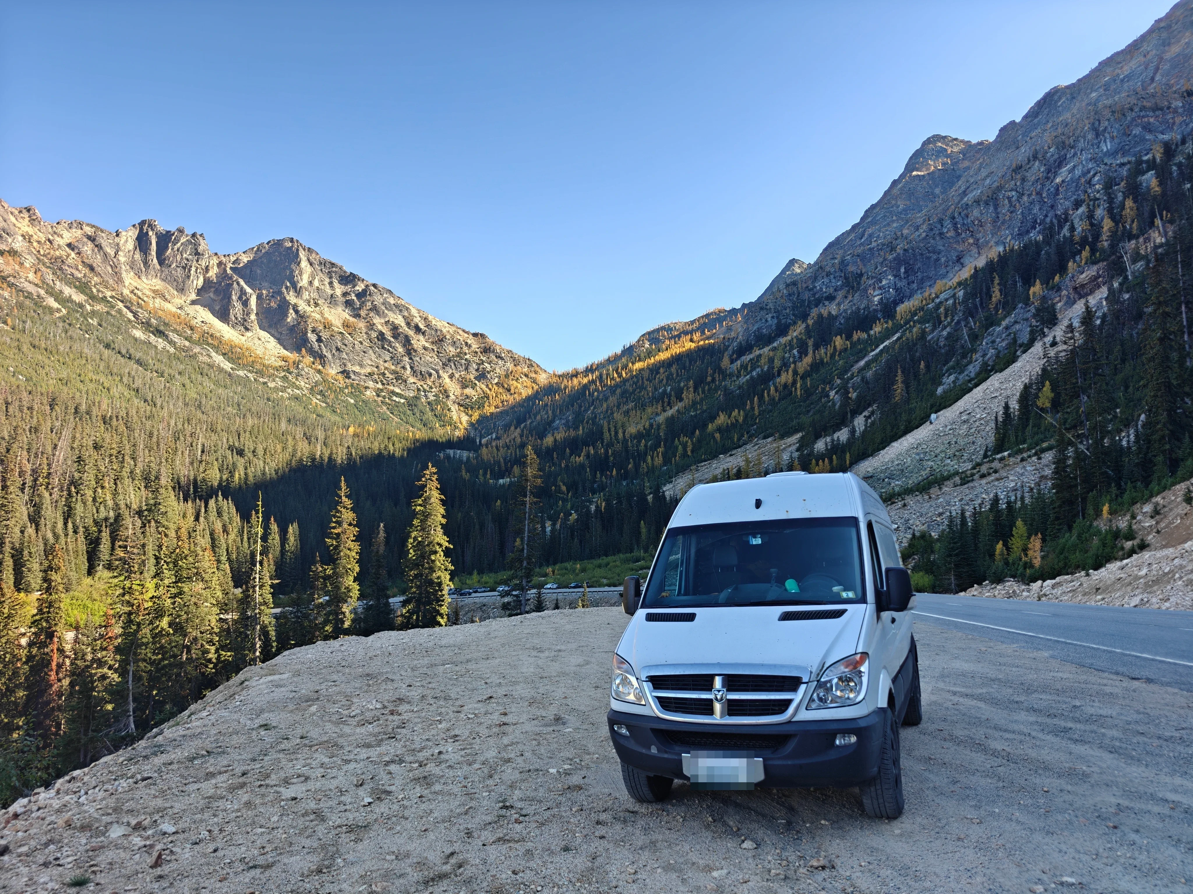 White camper van parked at a gravel highway pullout below larch-covered mountains