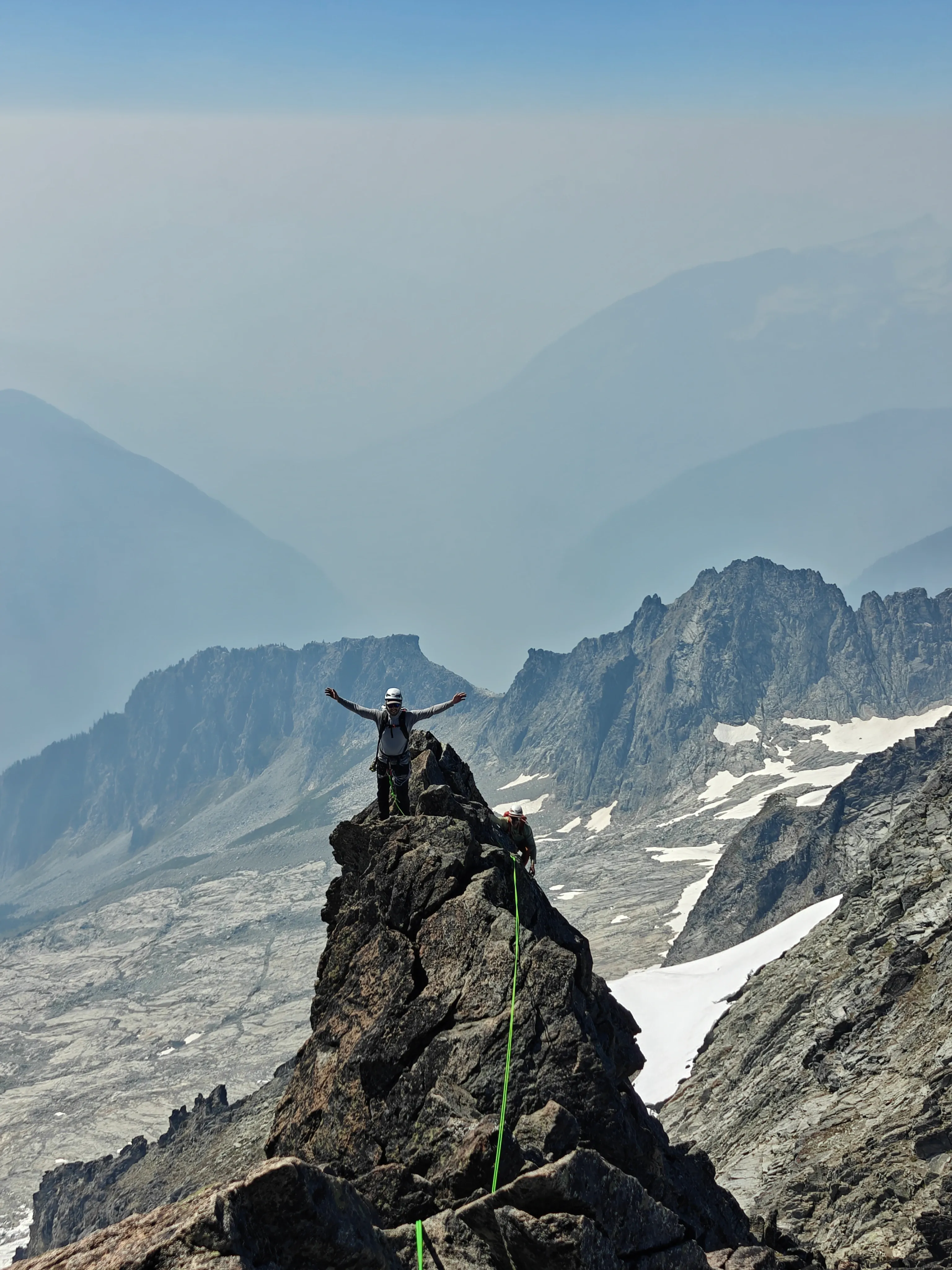 Climber standing with arms spread wide on top of a gendarme on the West Ridge of Forbidden Peak, rope trailing to a second climber below